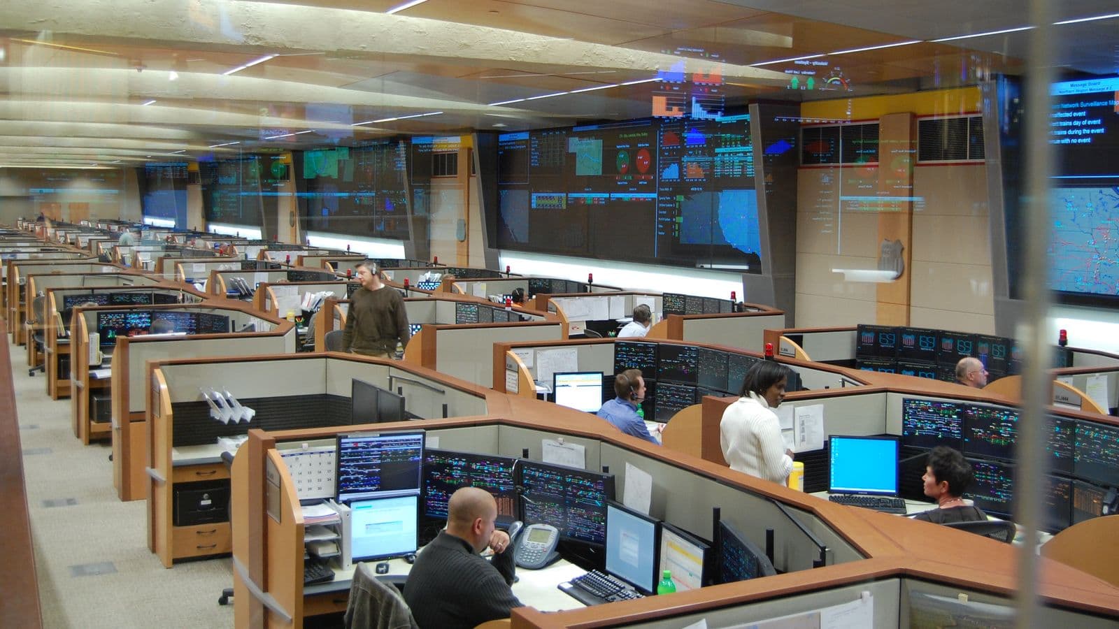 Dispatchers at desks with many monitors in a large U.S. railroad dispatching center, below wall-size network displays.