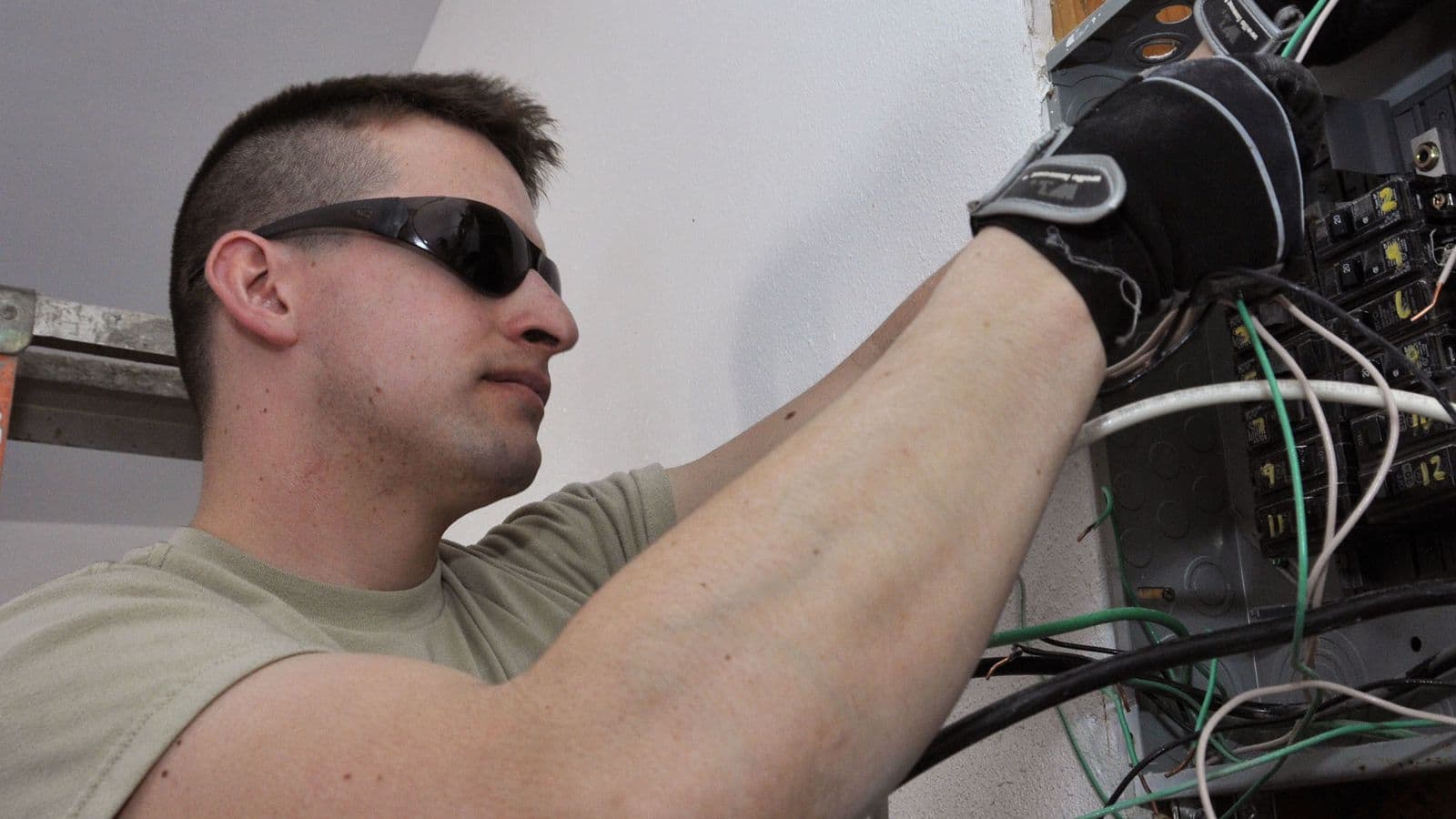 An electrician in safety glasses and gloves works on a crowded breaker panel during a panel upgrade in California.