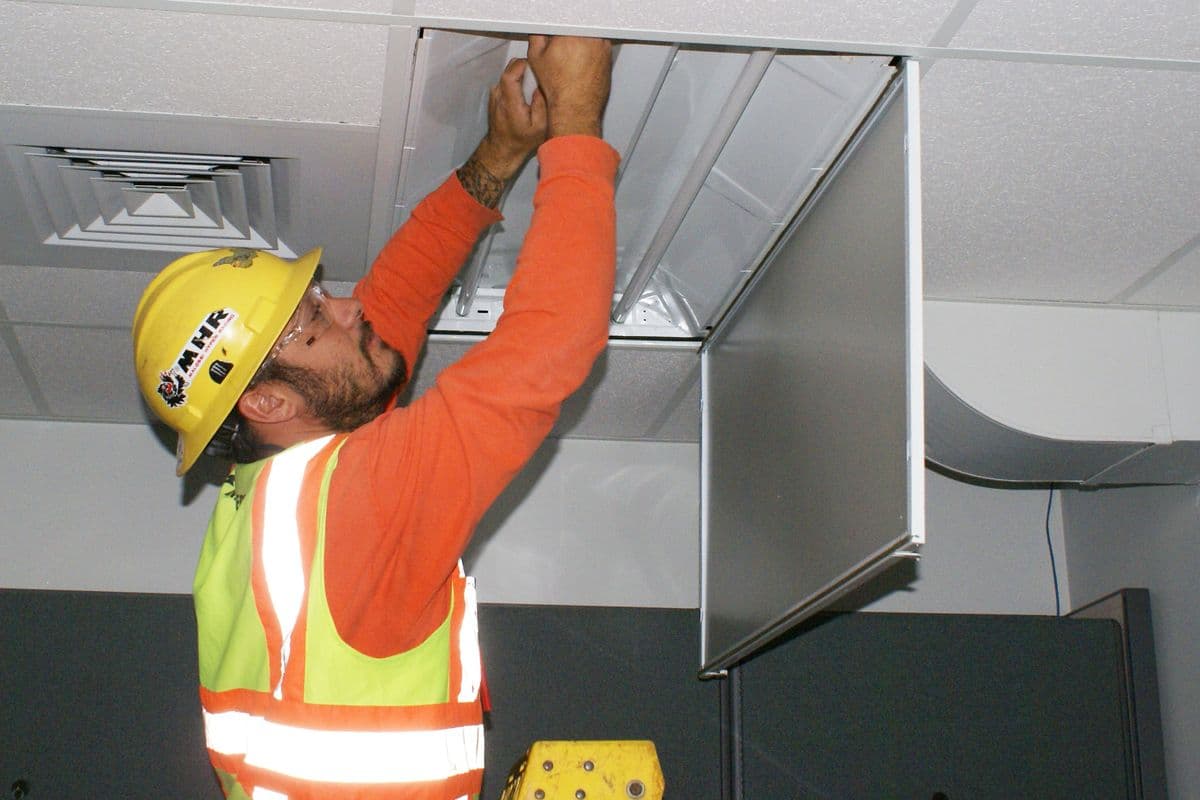An electrician in a hard hat and safety vest replaces light fixture ballasts in an office ceiling at Pearl Harbor, Hawaii.