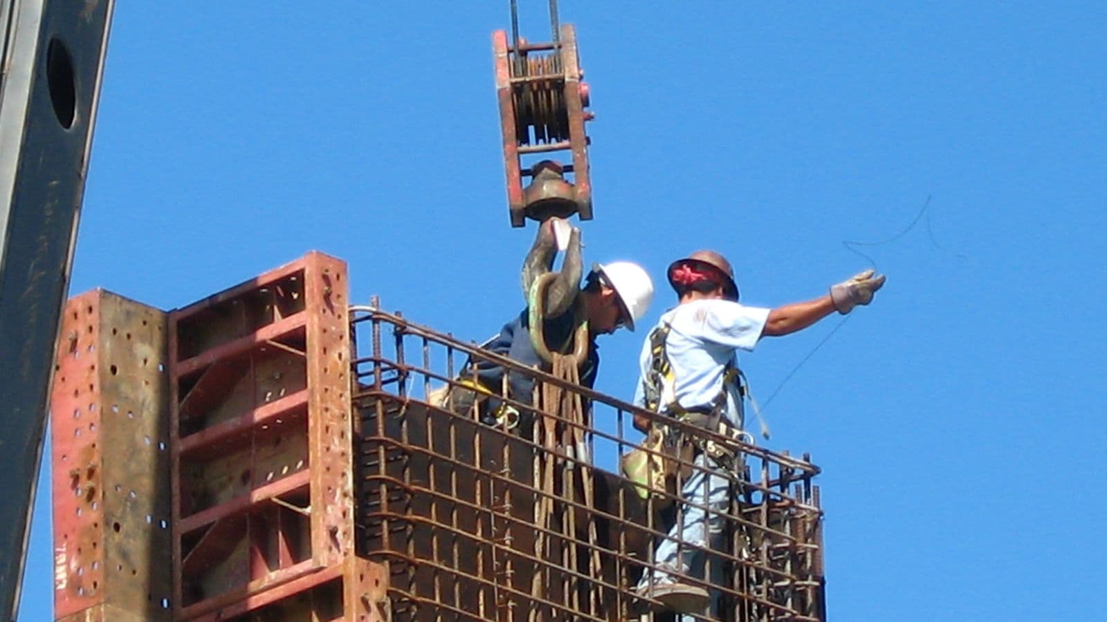 Two construction workers on top of a steel rebar cage guide a crane hook against a clear blue sky at a Houston, Texas, freeway project.