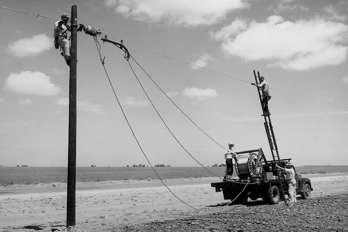 Black-and-white photo of a crew on a pole, a ladder and a truck with a cable reel, stringing lines across flat farmland.